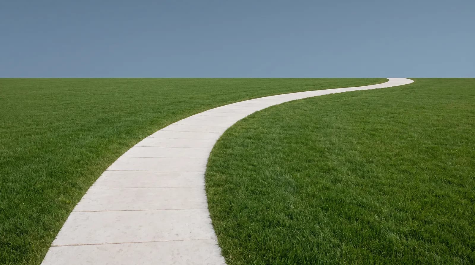 A white paved path curving away through flat green grass to the horizon under a clear sky