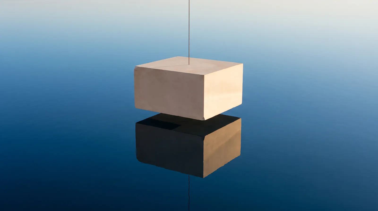 A pale block hanging from a single thin thread above still blue water, with its reflection below