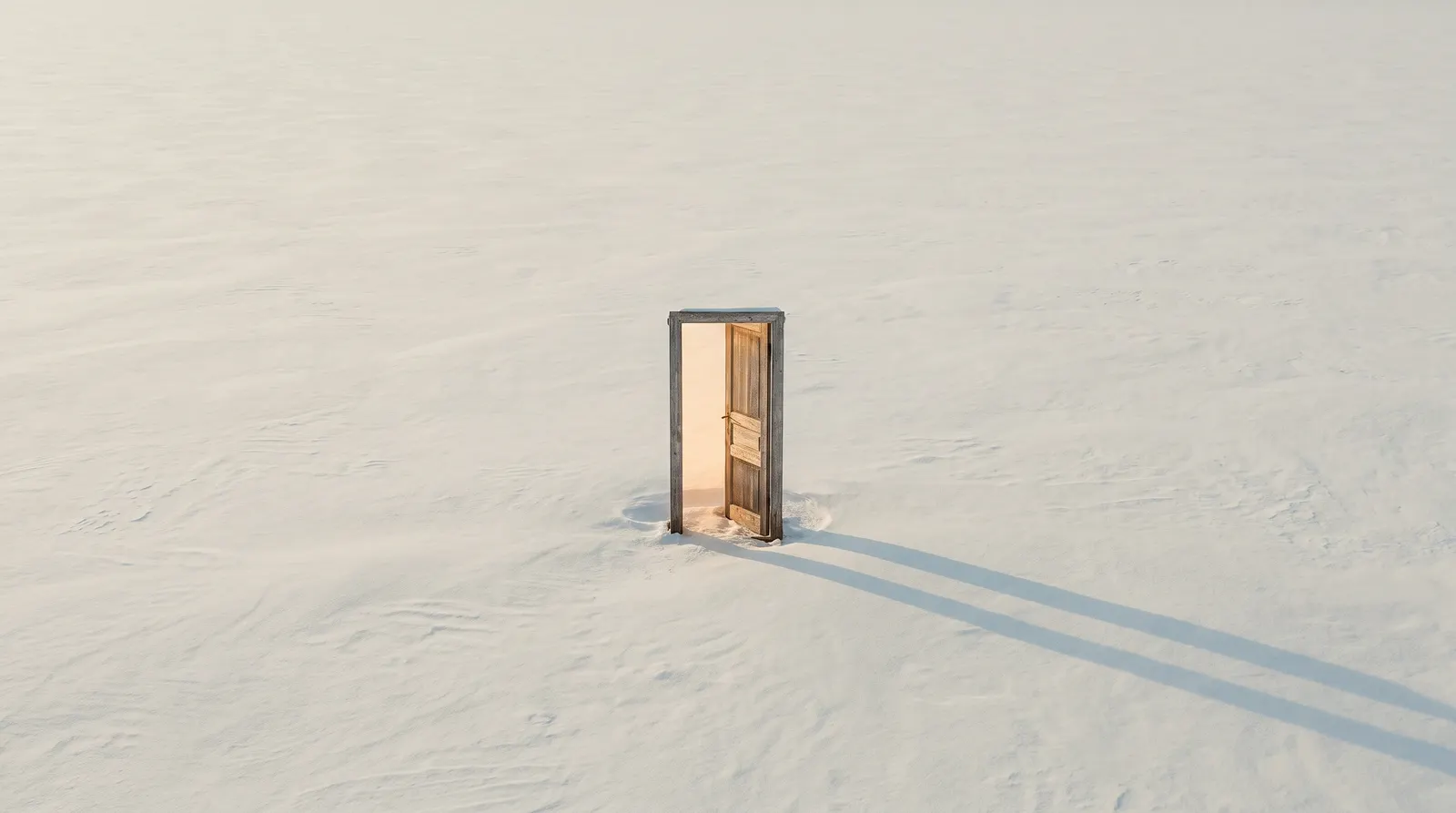 A weathered wooden door standing open in flat windblown snow, warm light in the gap and a long shadow behind it