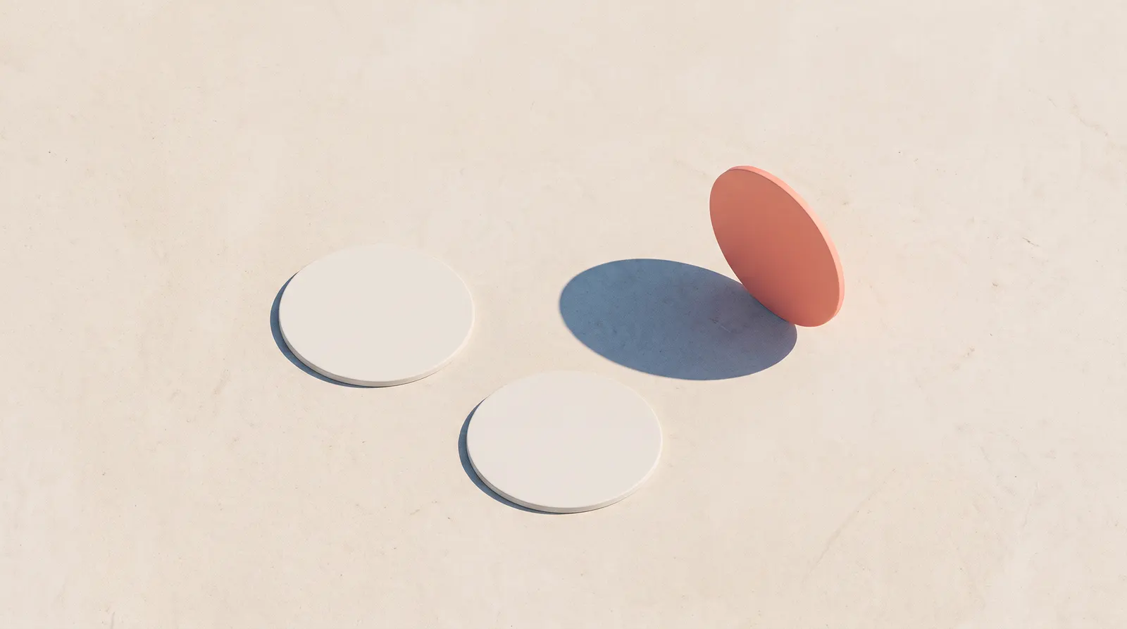 Three discs on warm sand, two lying flat and pale, one terracotta disc tipped up on its edge