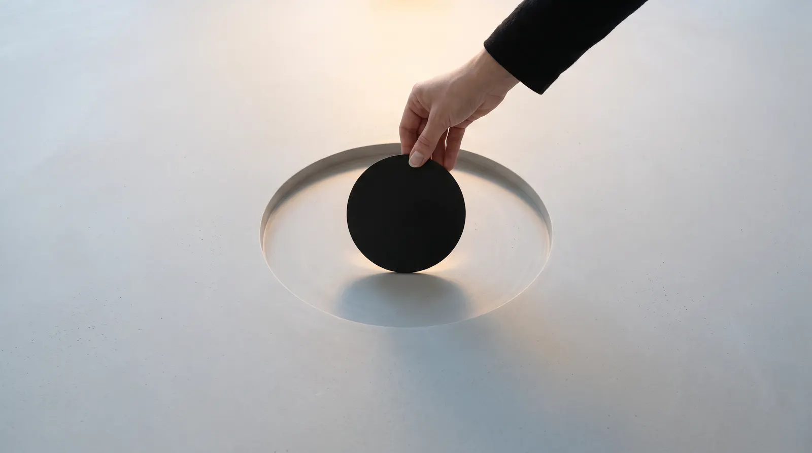 A hand in a dark sleeve lowering a black disc into a round opening in a white surface