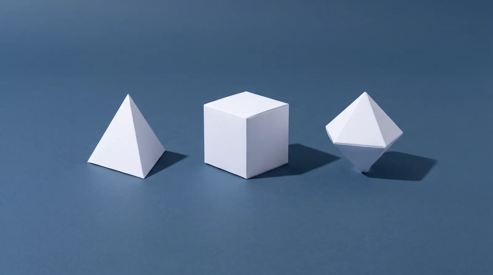 Three white paper forms on a blue-grey ground: a pyramid, a cube, and an octahedron resting on one point