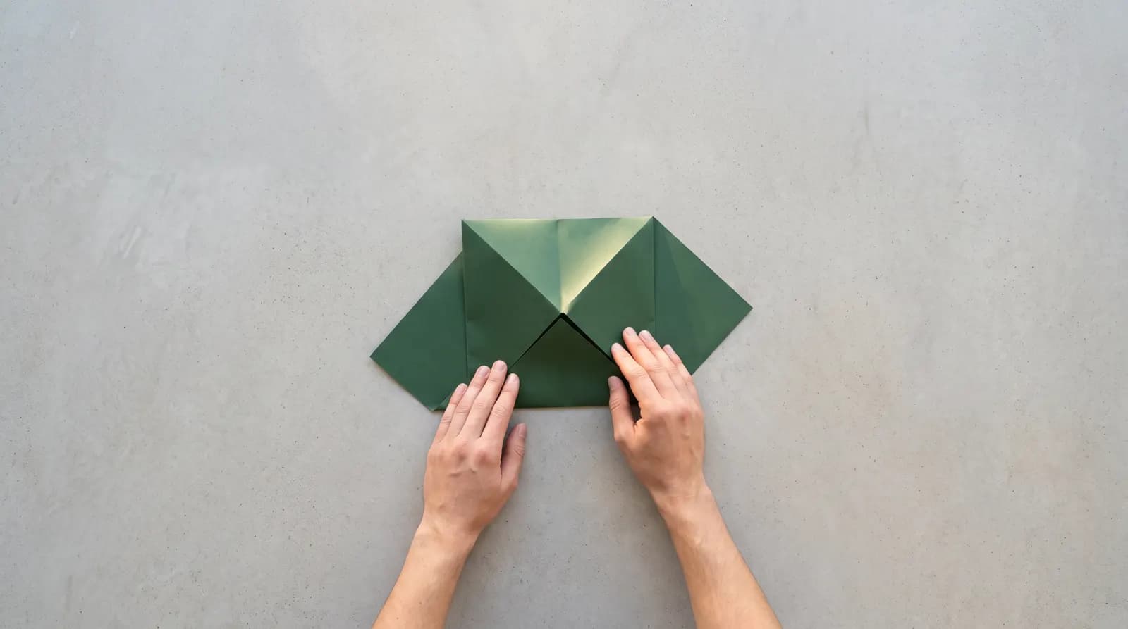 Two hands flattening a folded green paper fortune teller on a grey concrete surface